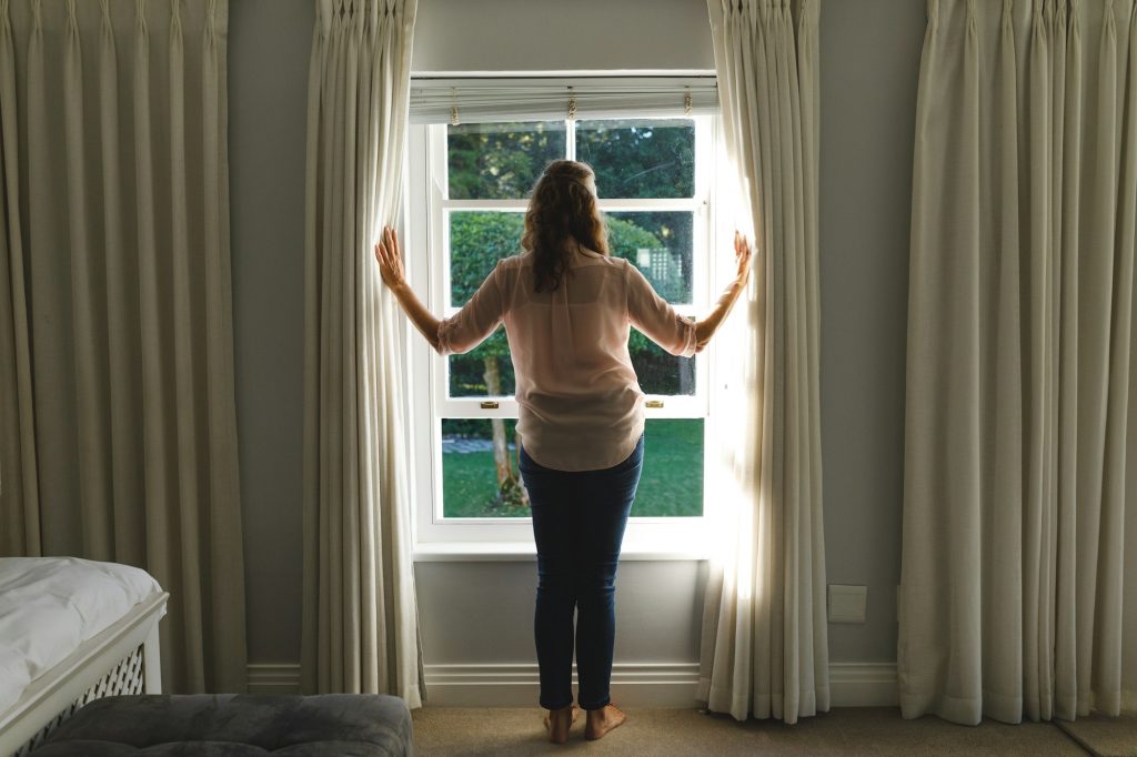 Thoughtful senior caucasian woman in bedroom, standing next to window, opening curtains
